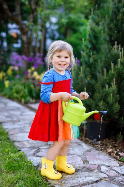 Krásné malé batole dívka ve žluté gumové boty a barevné šaty zalévání jarní květiny s dětmi vody může. Šťastné dítě pomáhá v rodinné zahradě, venku. — Stock fotografie