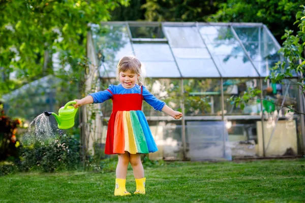 Wunderschönes kleines Mädchen in gelben Gummistiefeln und buntem Kleid, das Frühlingsblumen mit einer Wasserkanne gießt. Glückliches Kind hilft im Garten der Familie, im Freien. — Stockfoto