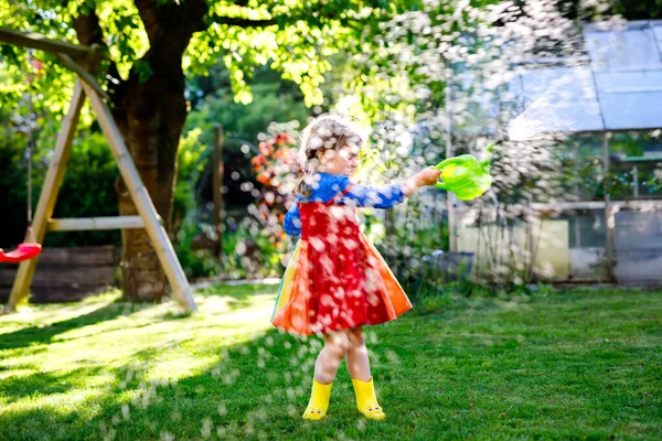 Bella bambina in stivali di gomma gialla e colorato vestito irrigazione fiori primaverili con acqua per bambini può. Bambino felice che aiuta nel giardino di famiglia, all'aperto, divertirsi con spruzzi — Foto Stock