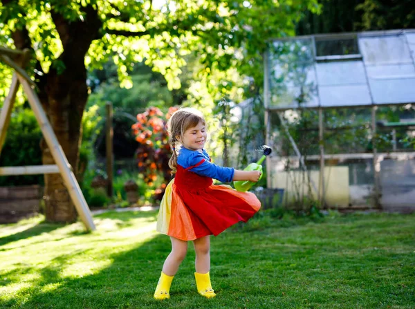 Beautiful little toddler girl in yellow rubber boots and colorful dress watering spring flowers with kids water can. Happy child helping in family garden, outdoors, having fun with splashing — Stock Photo, Image