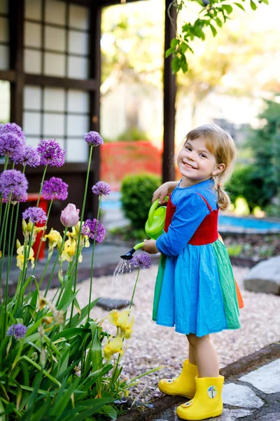 Wunderschönes kleines Mädchen in gelben Gummistiefeln und buntem Kleid, das Frühlingsblumen mit einer Wasserkanne gießt. Glückliches Kind hilft im Garten der Familie, im Freien. — Stockfoto