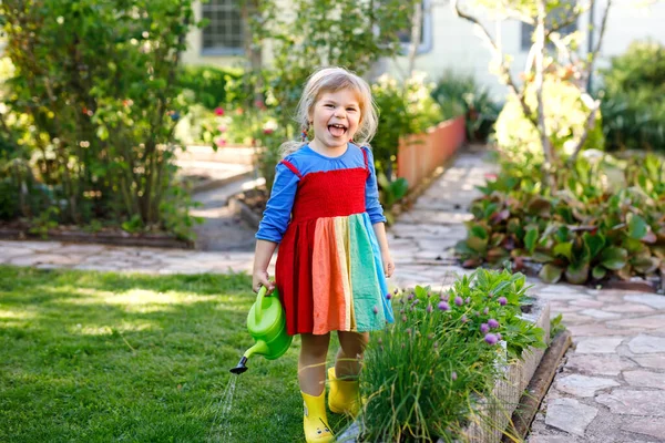 Beautiful little toddler girl in yellow rubber boots and colorful dress watering spring flowers with kids water can. Happy child helping in family garden, outdoors. — Stock Photo, Image