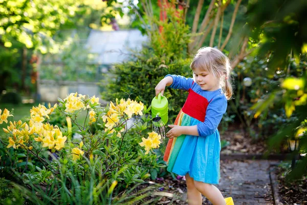 Piękna dziewczynka maluch w żółtych gumowych butach i kolorowe sukienki podlewanie wiosenne kwiaty z dziećmi wody może. Szczęśliwe dziecko pomaga w ogrodzie rodzinnym, na zewnątrz. — Zdjęcie stockowe