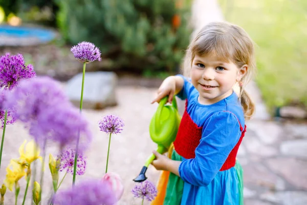 Piękna dziewczynka maluch w żółtych gumowych butach i kolorowe sukienki podlewanie wiosenne kwiaty z dziećmi wody może. Szczęśliwe dziecko pomaga w ogrodzie rodzinnym, na zewnątrz. — Zdjęcie stockowe