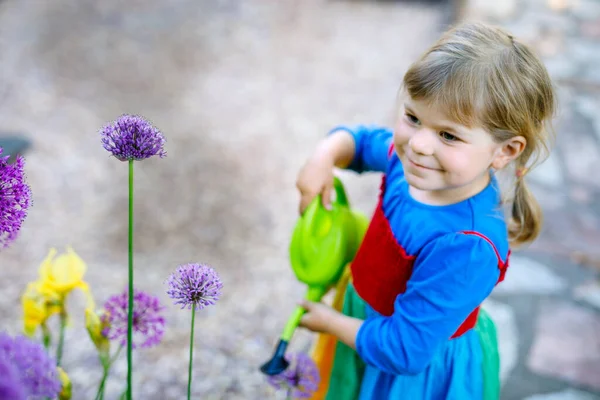 Beautiful little toddler girl in yellow rubber boots and colorful dress watering spring flowers with kids water can. Happy child helping in family garden, outdoors. — Stock Photo, Image