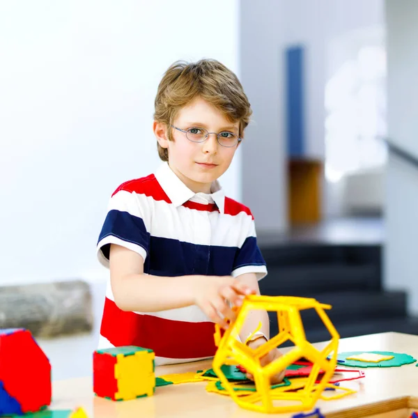 Маленький мальчик в очках играет с пластмассовым набором элементов в школе или детском саду. Счастливый ребенок строит и создает геометрические фигуры, учится игре и геометрии. — стоковое фото