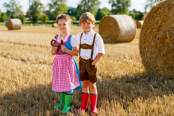 Dvě děti v tradičních bavorských kostýmech na pšeničném poli. Během Oktoberfestu jedly německé děti chléb a preclík. Chlapec a dívka hrát na balíky sena během letní sklizně v Německu. — Stock fotografie