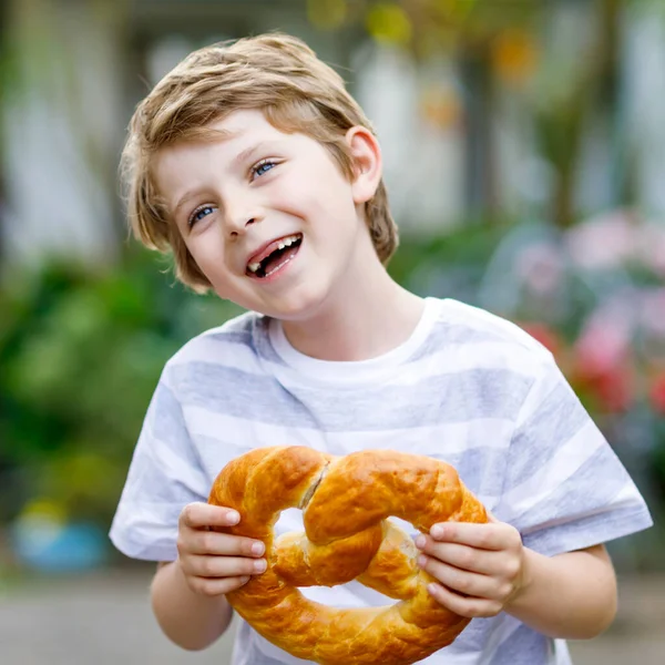 Очаровательный маленький мальчик ест огромный баварский крендель. Счастливый блондин с разницей в зубах наслаждается со вкусом хлебом. Здоровое питание для счастливых детей . — стоковое фото