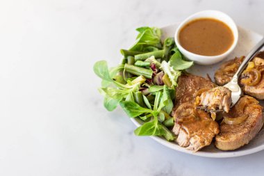 Kızarmış domuz pirzolası, karamelize soğan ve sosla birlikte yeşil salata ve fasulye ile servis ediliyor. Açık gri arkaplan, seçici odak.