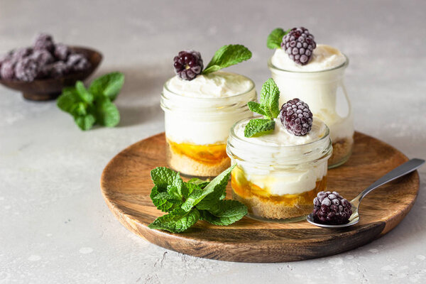 Homemade no baked dessert (cheesecake, trifle, mousse) with orange or peach jam in small portioned jars served with frozen berries and mint. Light grey stone background, copy space.