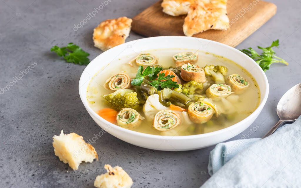 Soup with vegetables and egg pancakes in a white plate, light grey background. Traditional German Flaedlesuppe and Austrian Frittatensuppe. 