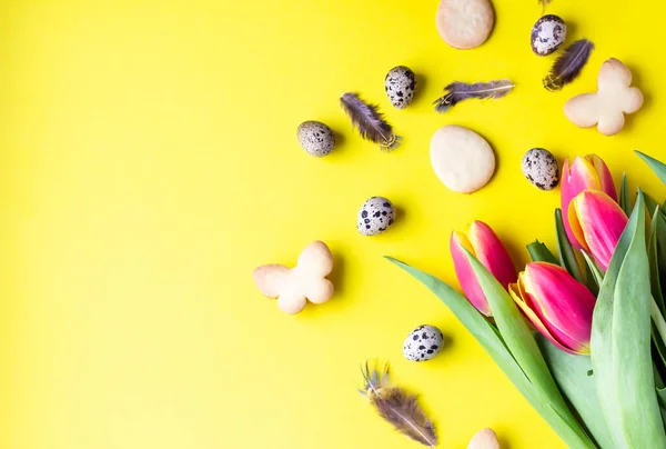Easter cookies, red and yellow tulips, quail eggs on yellow paper background. Easter symbols and traditions. Butter and sugar cookies.