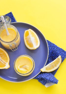 Mavi seramik tabakta iki kavanoz ev yapımı limon, krema ve taze limon. Sarı kağıt arka plan. Seçici odak.