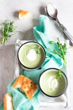 Tahta tepside servis edilen taze Focaccia ve mikro yeşilliklerle kremalı sağlıklı brokoli çorbası, açık gri beton arka plan. Üst görünüm.