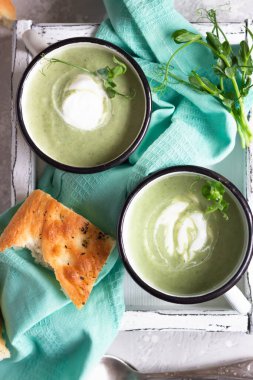 Tahta tepside servis edilen taze Focaccia ve mikro yeşilliklerle kremalı sağlıklı brokoli çorbası, açık gri beton arka plan. Üst görünüm.