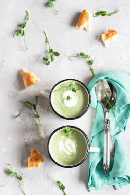 Lezzetli brokolili krema çorbası, taze ekmek ve açık gri beton arka planda mikro yeşilliklerle servis edilmiş bir kupa. Üst görünüm.