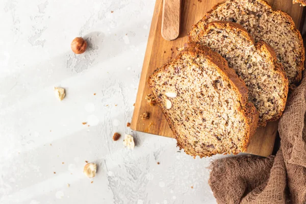 Homemade banana bread or cake with hazelnut on wooden cutting board, light stone background. Trendy soft shadows, atmospheric lighting, selective focus. Flat lay composition.