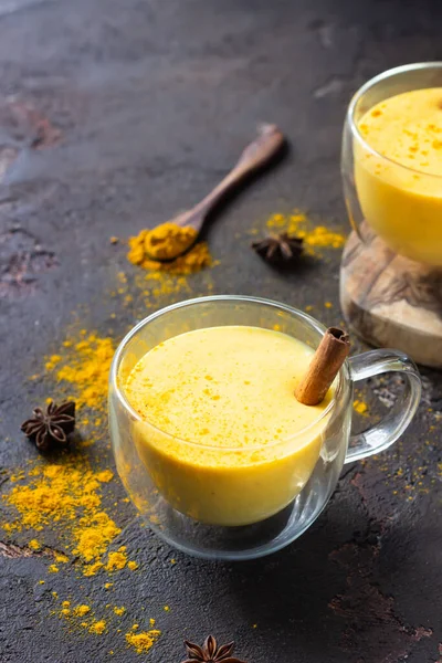 Golden milk or latte with turmeric (curcuma) powder with spices, dark brown concrete background. Trendy detox, immune boosting, anti-inflammatory healthy cozy drink.