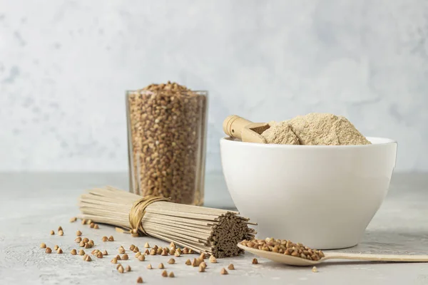 Bowl with gluten free buckwheat flour, buckwheat grains in a glass and soba noodles. Light grey concrete background. Healthy diet.