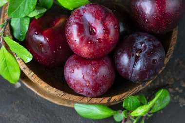 Tahta kasede yaprakları ve koyu beton arka planı olan olgun, sulu mor erikler. Kapat..