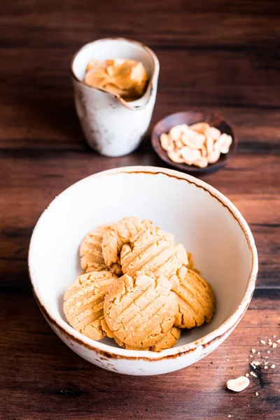 Heap of homemade peanut butter cookies in a bowl with a plate of peanuts and a cup of peanut butter on a wooden table, selective focus. Image with copy space. Traditional american treat.