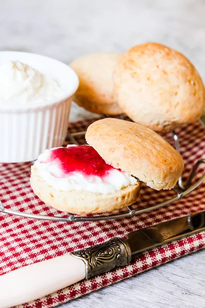 Traditional british pastries or sweet buns called scones with vanilla cream cheese and fresh strawberry jam on a cooling rack on a wooden table, selective focus. Breakfast, snack, picnic food.