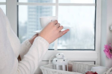 Bir fincan sıcak çay ya da kahve içen bir kadının ellerini kapayın ve sabahları evdeki pencereye bakın. Boşluğu metin olarak kopyala, karantina konseptinde güvenli bir şekilde evde kal.