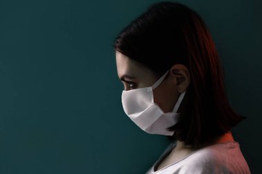 Tıbbi maskeli izole bir kız profili yakın plan, dramatik bir portre, duvara dayalı, kırmızı arka plan ışığı