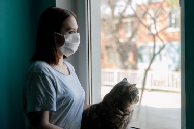 Evin penceresinde kedisiyle birlikte maskeli bir kız.