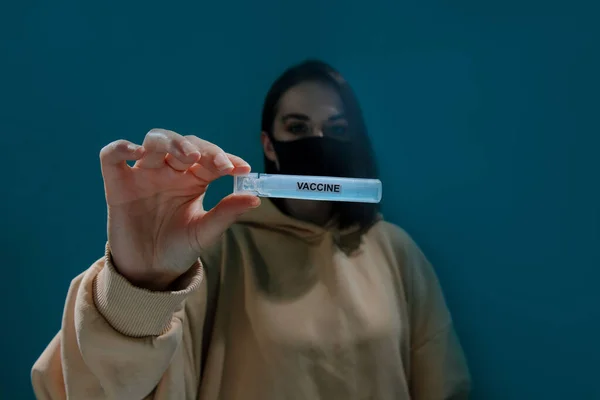 A fashionable girl in a black medical mask and beige hoodie holds a test tube with a vaccine in her hand