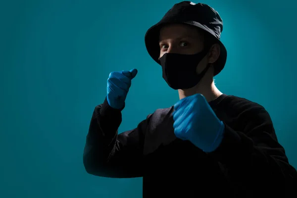 A young guy in a black medical mask in a black bucket hat, in a black jacket in blue medical gloves holds his fists, medical blue gloves