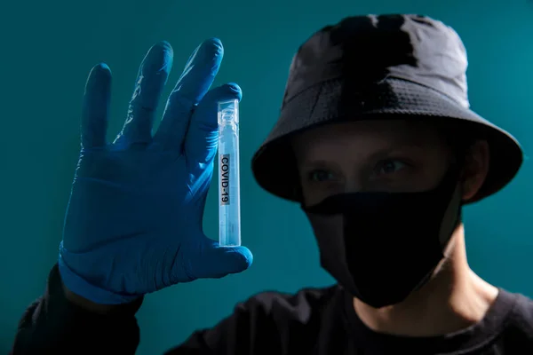 A guy in a black medical mask, a black bucket hat, a black jacket, and blue medical gloves is holding a flask with a virus