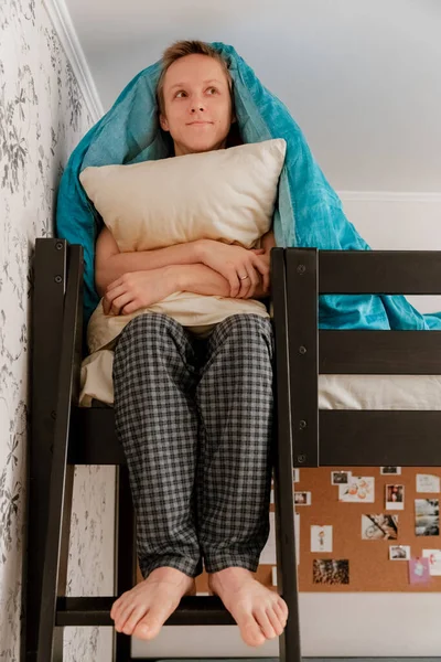 The guy is sitting on a bunk bed under a blue blanket and hugs a pillow, dreamy, vertically