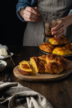 Kadın elleri taze pişmiş brioche 'nin üzerine bal döküyor. Çikolata parçacıkları, tereyağı, siyah ahşap arka planda süt.
