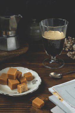 Kahve bardağına yakın plan, biraz karamelli şekerleme, koyu ahşap masa üzerinde moka kahve makinesi.