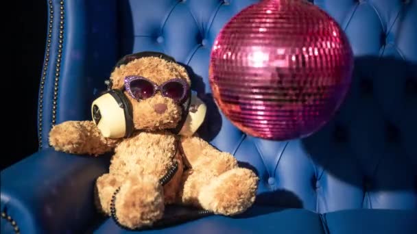 棕色泰迪熊绒毛玩具 戴着太阳镜和耳机 沙发背景是迪斯科球 — 图库视频影像