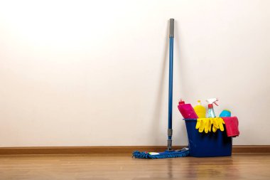 İçinde temizlik ürünleri ve deterjanlar olan çeşitli şişeler, mavi kovadaki bez ve yerdeki paspas.