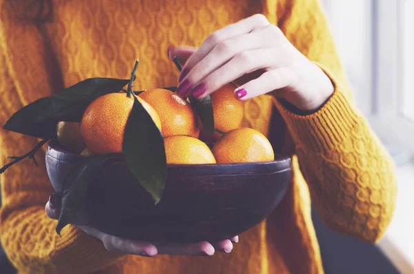 Chica en casa sosteniendo cuenco de madera lleno de mandarinas de invierno maduras —  Fotos de Stock