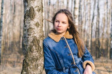 Uzun saçlı, uzun saçlı genç bir kız bir ağaca yaslanır ve güneşli bir huş ağacının dibine bakar.