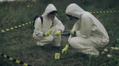 Bakteriyolojik koruma ekipmanlarıyla iki kişi açık havada cesedin yanındaki plastik torbada kanıt topluyor.