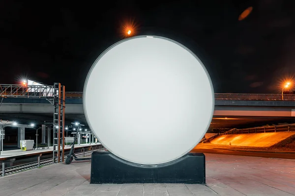 Blank round white outdoor banner stand next to bright modern of railway metro station. night time