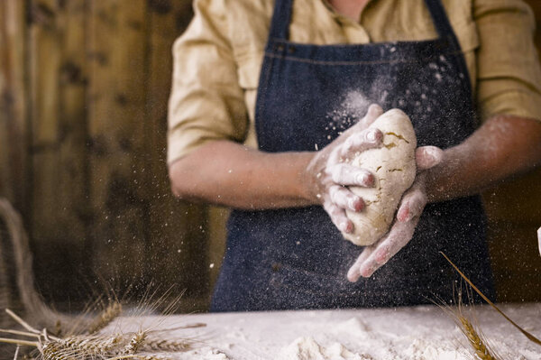 Womens hands, flour and dough. Levitation in the frame. A woman in an apron is preparing dough for home baking. Rustic style photo. Wooden table, wheat ears and flour in the crucifix. Emotional photo