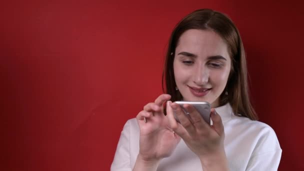 Una giovane ragazza tiene in mano un telefono. Girare su uno sfondo rosso in studio. Comunicazione smartphone e tecnologia moderna. — Video Stock