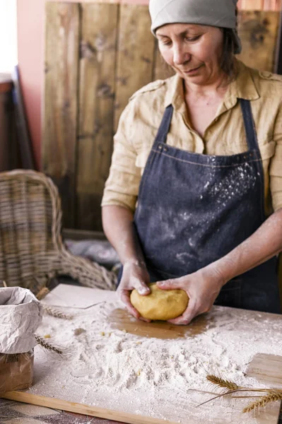 Kvinnliga händer och deg. En kvinna i förkläde förbereder degen för hembakning. Rustik stil foto. Träbord, veteöron och mjöl. Fritt utrymme för text. Känslomässigt foto. — Stockfoto