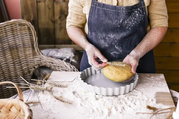 Manos femeninas, harina, masa y una sartén para pasteles. Una mujer en un delantal está preparando masa para hornear en casa. Foto de estilo rústico. Mesa de madera, espigas de trigo y harina. Foto emocional. Ambiente acogedor. — Foto de Stock