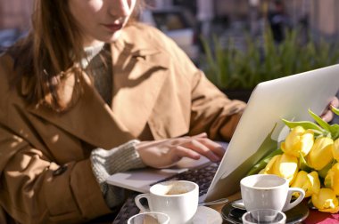 Genç bir kız caddedeki bir kafede kahve içiyor ve internette dizüstü bilgisayarla çalışıyor. Çerçevede güneş ışığı. Noutbuk 'la seyahat edip yürümek. Boşluğu kopyala.