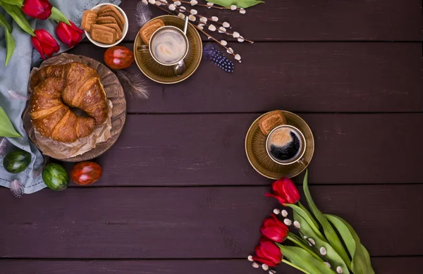 Paskalya kahvaltısında aromatik taze espresso, kruvasan, renkli yumurta, kırmızı lale ve söğüt. Kurabiyeli kahve, ahşap masadaki çiçekler. Yukarıdan bak. Boşluğu kopyala. — Stok fotoğraf