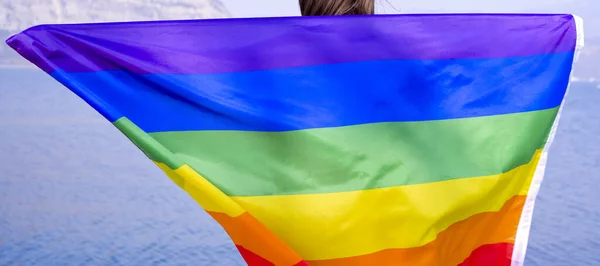 Mujer sosteniendo bandera arco iris gay cerca del lago, montañas al aire libre. El concepto de felicidad, libertad y amor para parejas del mismo sexo. Copiar espacio. — Foto de Stock