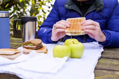 Parkta eski ahşap bir masada öğle yemeği. Termosta sandviç, elma ve kahve. Çerçevedeki ağaçların arasından güneş ışığı. Bahar