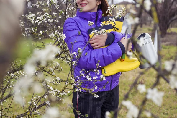 Una ragazza in una giacca viola tiene stivali di gomma giallo brillante, annaffiatoio, una vanga nelle sue mani. Il periodo di fioritura degli alberi nei giardini e l'orario di lavoro su appezzamenti personali. Settori sfocati — Foto Stock